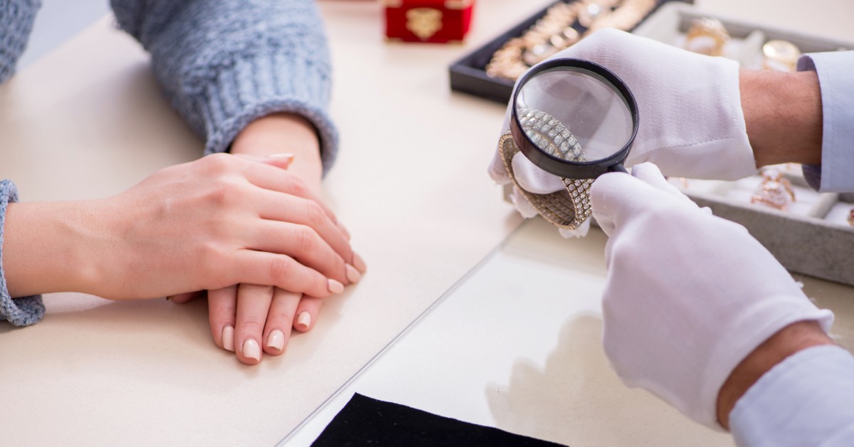 A pair of hands wearing gloves looking at a diamond bracelet with a magnifying glass in front of another pair of hands.