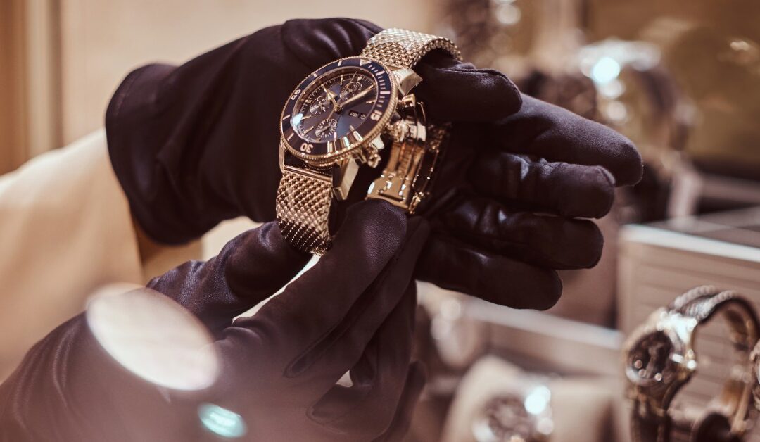 Two hands wearing black gloves, holding a gold watch with a black frame, with more watches in the background.